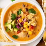 A bowl of creamy chicken taco soup topped with avocado, red onion, cilantro, and crumbled tortilla chips. It’s set on a wooden surface with tortilla chips on the side.