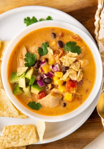 A bowl of creamy chicken taco soup topped with avocado, red onion, cilantro, and crumbled tortilla chips. It’s set on a wooden surface with tortilla chips on the side.