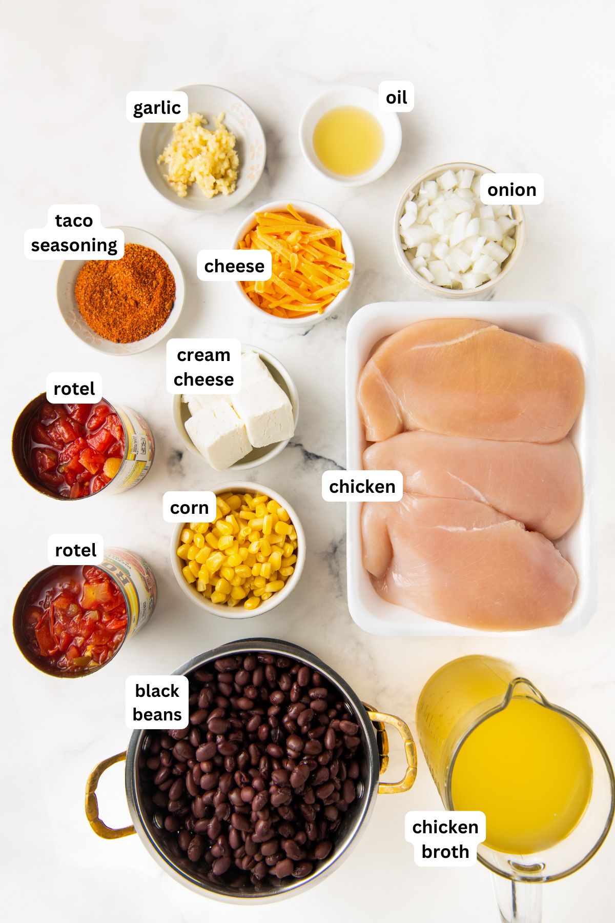Ingredients for chicken taco soup recipe laid out on a white surface: raw chicken, diced onion, garlic, oil, taco seasoning, shredded cheese, cream cheese, rotel tomatoes, corn, black beans, and chicken broth.