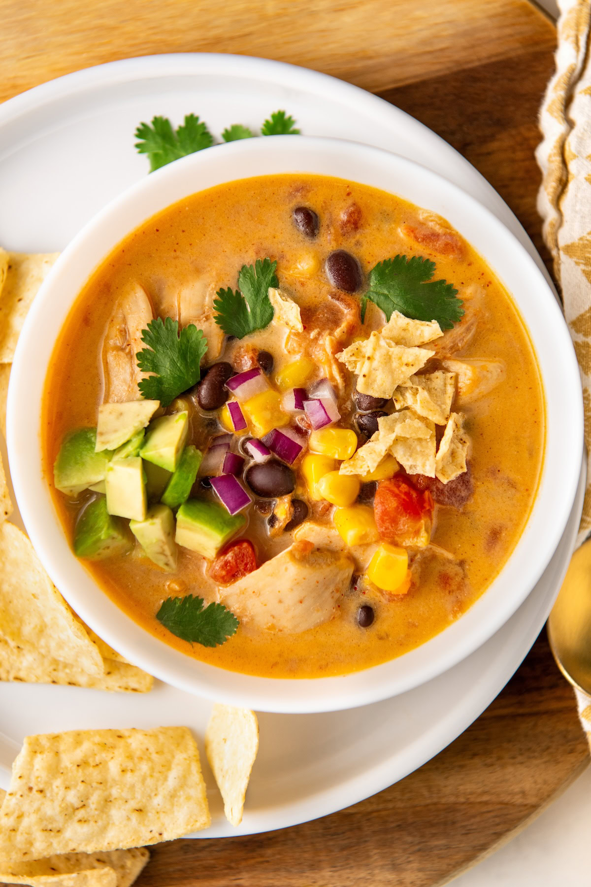 Made quickly and easily in just one-pot, this creamy Chicken Taco Soup recipe is comforting, hearty, and packed with bold Tex-Mex flavors! A bowl of creamy chicken taco soup topped with avocado, red onion, cilantro, and crumbled tortilla chips. It’s set on a wooden surface with tortilla chips on the side.