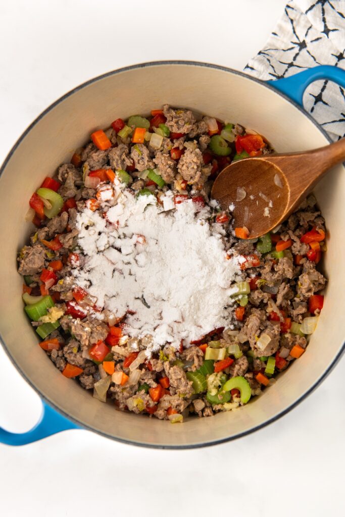 A blue pot filled with a mixture of browned Italian sausage, chopped red bell pepper, celery, garlic and onions. A wooden spoon rests inside, and a pile of white flour is on top of the mixture, ready to be stirred in.