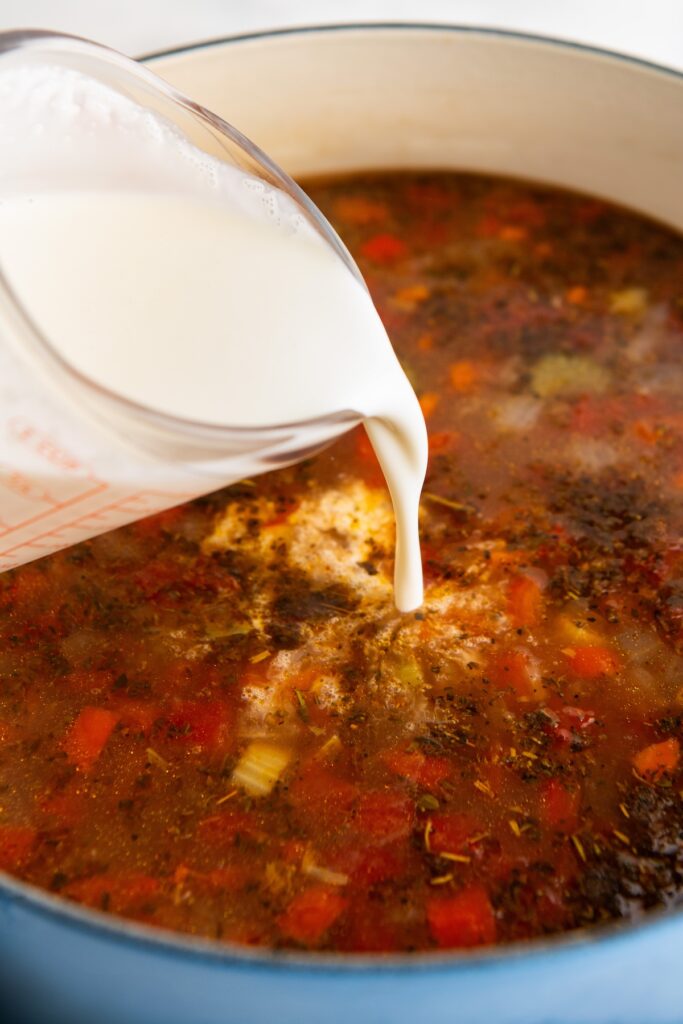 Close-up of a measuring cup pouring cream into a pot of soup. The soup, filled with chopped vegetables and herbs, is simmering in a blue pot.