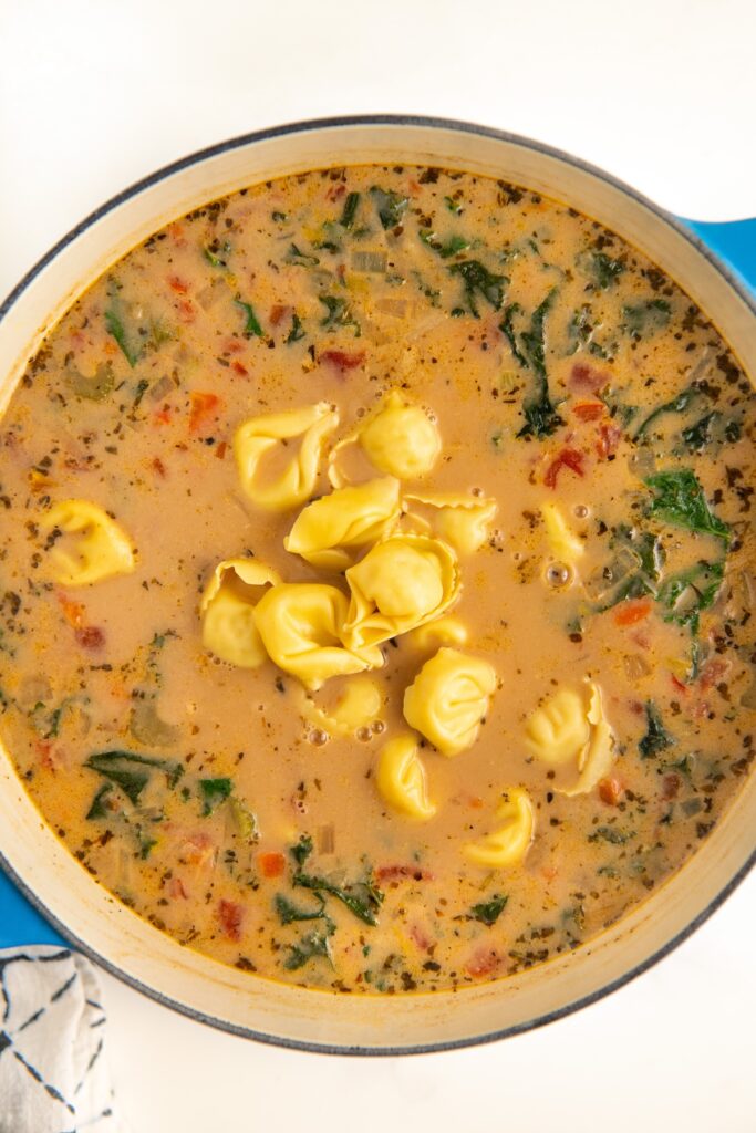 A blue pot filled with creamy tortellini sausage soup, featuring pasta, kale, and tomatoes in a rich, seasoned broth.