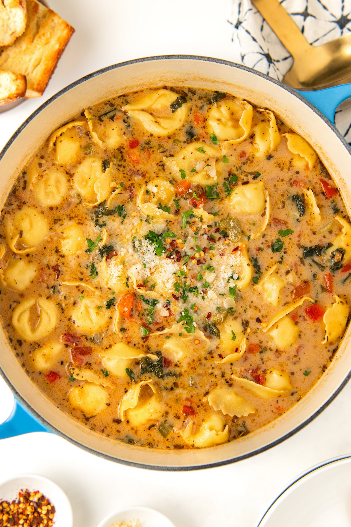 A large pot of creamy sausage and tortellini soup with various herbs and spices, featuring cheese-filled tortellini, diced tomatoes, and kale. A sprinkle of grated cheese and red pepper flakes is on top. Bread and utensils are nearby.
