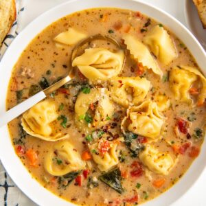 A bowl of creamy Italian sausage tortellini soup filled with kale, carrots, and diced tomatoes. Sprinkled with herbs and grated cheese, the soup is accompanied by a spoon and a small dish of Parmesan cheese on the side.