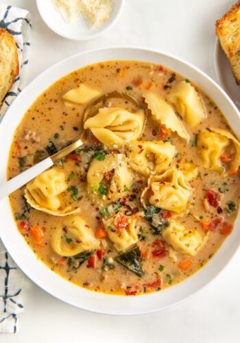 A bowl of creamy Italian sausage tortellini soup filled with kale, carrots, and diced tomatoes. Sprinkled with herbs and grated cheese, the soup is accompanied by a spoon and a small dish of Parmesan cheese on the side.