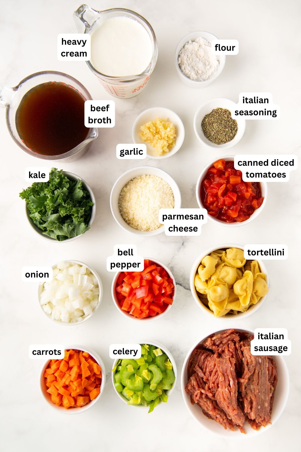 Ingredients for sausage tortellini soup recipe laid out on a white surface labeled clockwise: heavy cream in a measuring cup, flour, Italian seasoning, canned diced tomatoes, tortellini, Italian sausage, green bell pepper, celery, carrots, onion, bell pepper, kale, garlic, parmesan, beef broth.