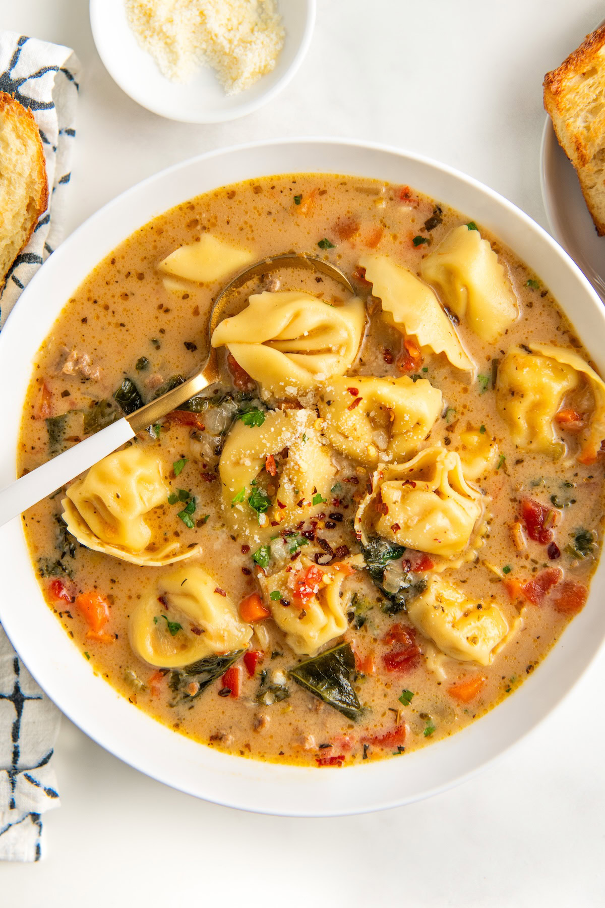 With Italian sausage, cheese tortellini, and fresh kale, this Creamy Sausage Tortellini Soup recipe is a one-pot dinner ready in 30 minutes! A bowl of creamy Italian sausage tortellini soup filled with kale, carrots, and diced tomatoes. Sprinkled with herbs and grated cheese, the soup is accompanied by a spoon and a small dish of Parmesan cheese on the side.