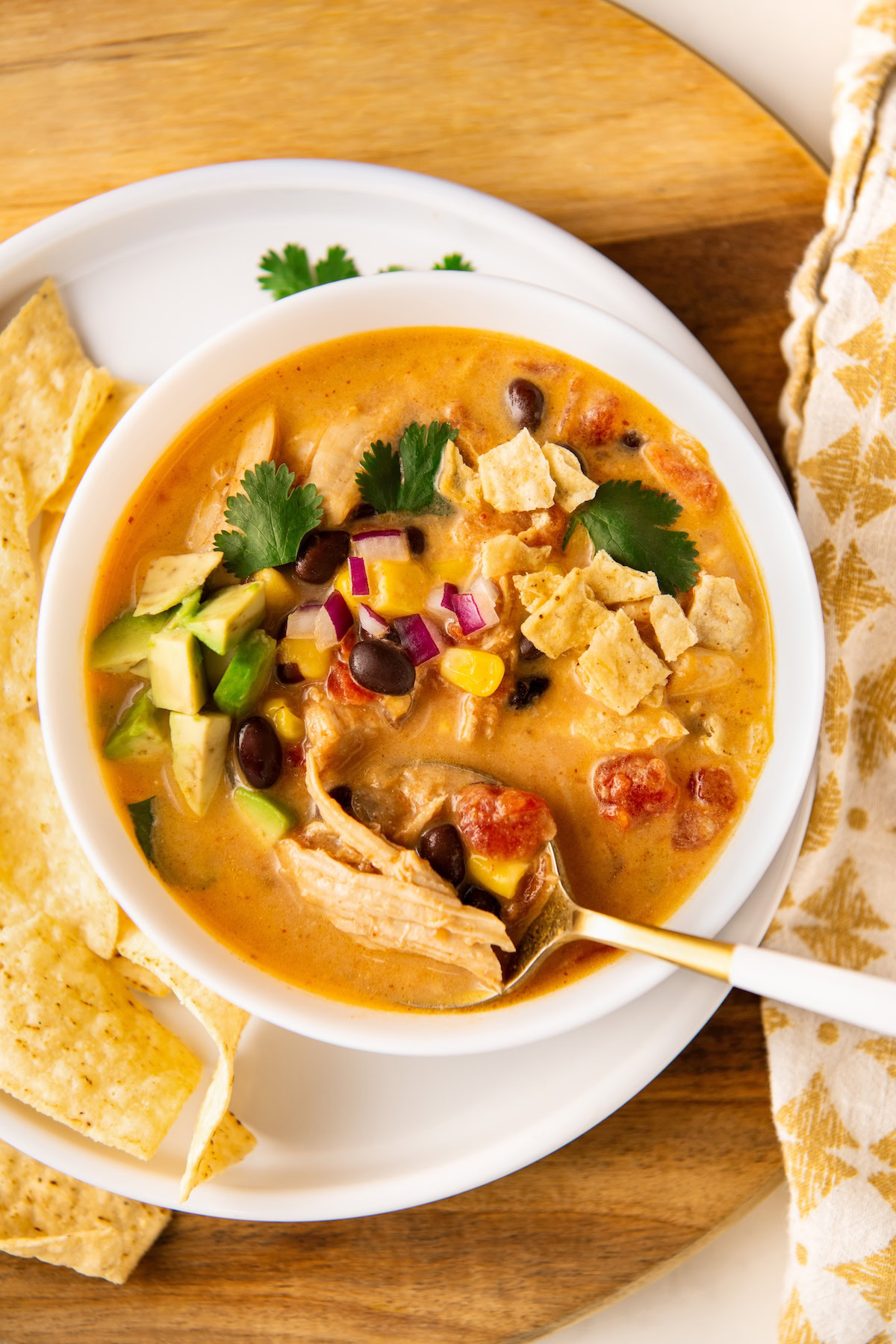 A bowl of creamy taco chicken soup garnished with avocado, tortilla chips, cilantro, and diced red onions on a white plate. A spoon is resting in the bowl, with tortilla chips on the side.