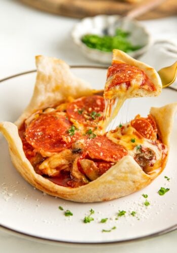 An upside-down pizza pot pie with pepperoni, cheese, and herbs sits on a white plate. A fork lifts a cheesy slice, showing melted cheese stretching. Chopped parsley and grated cheese garnish the pizza and plate.