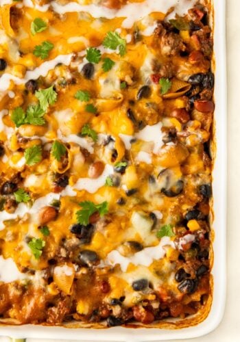 A close-up of a baked taco pasta casserole topped with melted cheese, black beans, corn, tomatoes, and fresh cilantro, with a drizzle of white sauce in a white baking dish.