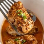 Crockpot cajun chicken breast is lifted with a spatula from a slow cooker filled with more chicken breasts and a flavorful orange-colored broth, garnished with chopped green onions.