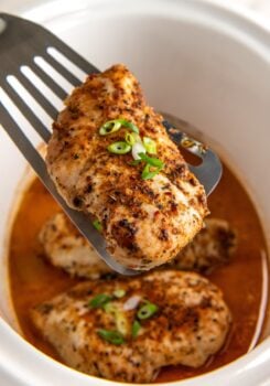 Crockpot cajun chicken breast is lifted with a spatula from a slow cooker filled with more chicken breasts and a flavorful orange-colored broth, garnished with chopped green onions.