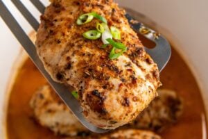 Crockpot cajun chicken breast is lifted with a spatula from a slow cooker filled with more chicken breasts and a flavorful orange-colored broth, garnished with chopped green onions.