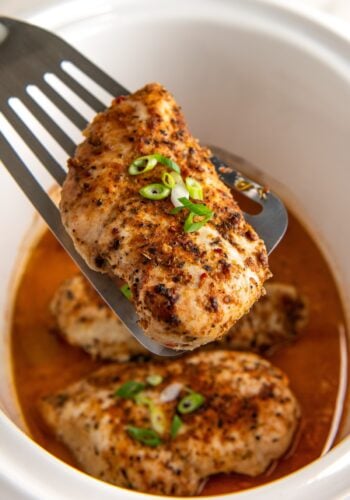 Crockpot cajun chicken breast is lifted with a spatula from a slow cooker filled with more chicken breasts and a flavorful orange-colored broth, garnished with chopped green onions.