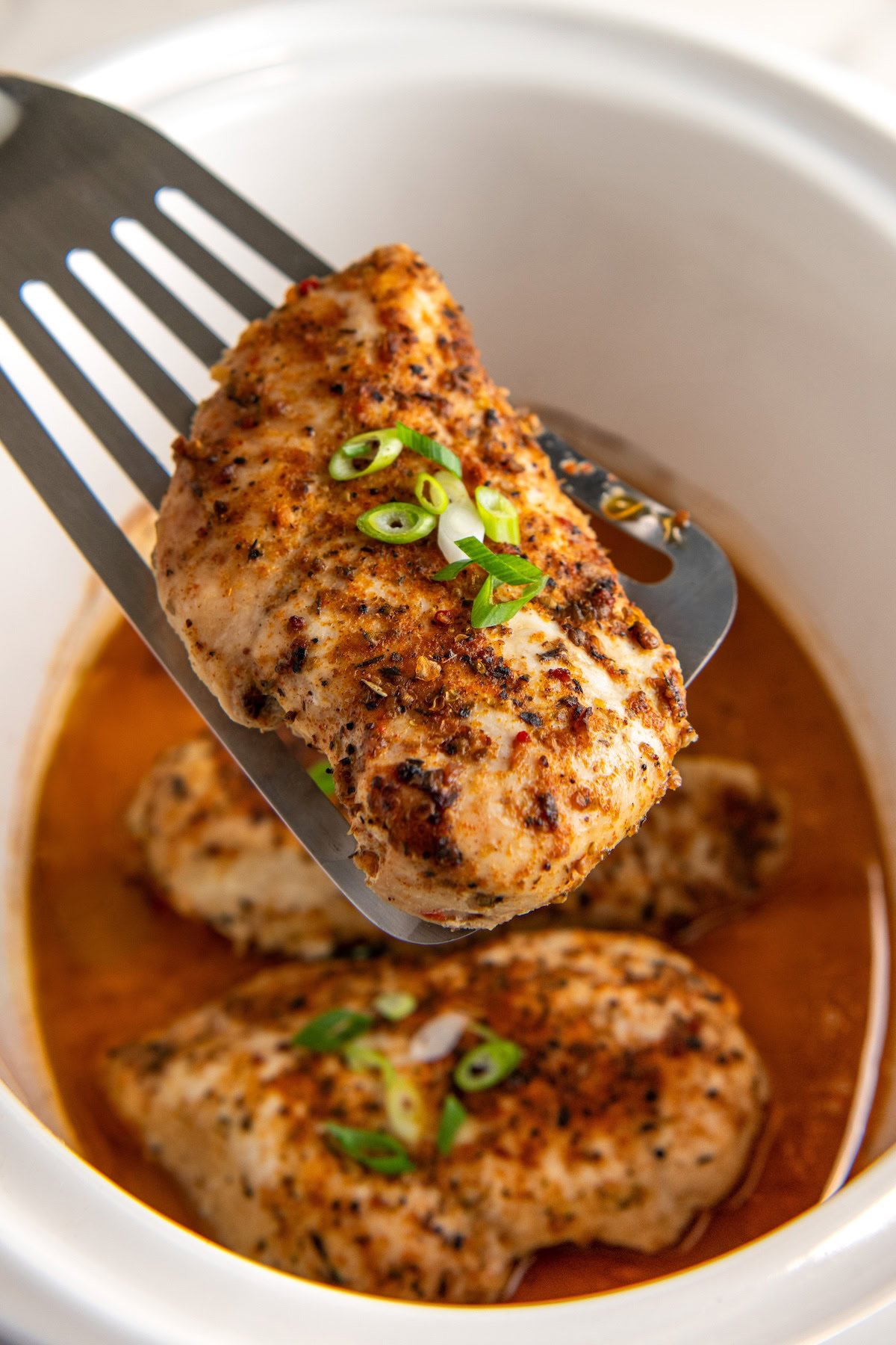 Crockpot cajun chicken breast is lifted with a spatula from a slow cooker filled with more chicken breasts and a flavorful orange-colored broth, garnished with chopped green onions.