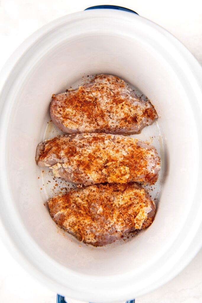 Three raw chicken breasts coated with butter and seasoned with cajun seasoning are arranged in a white slow cooker, ready to be cooked.