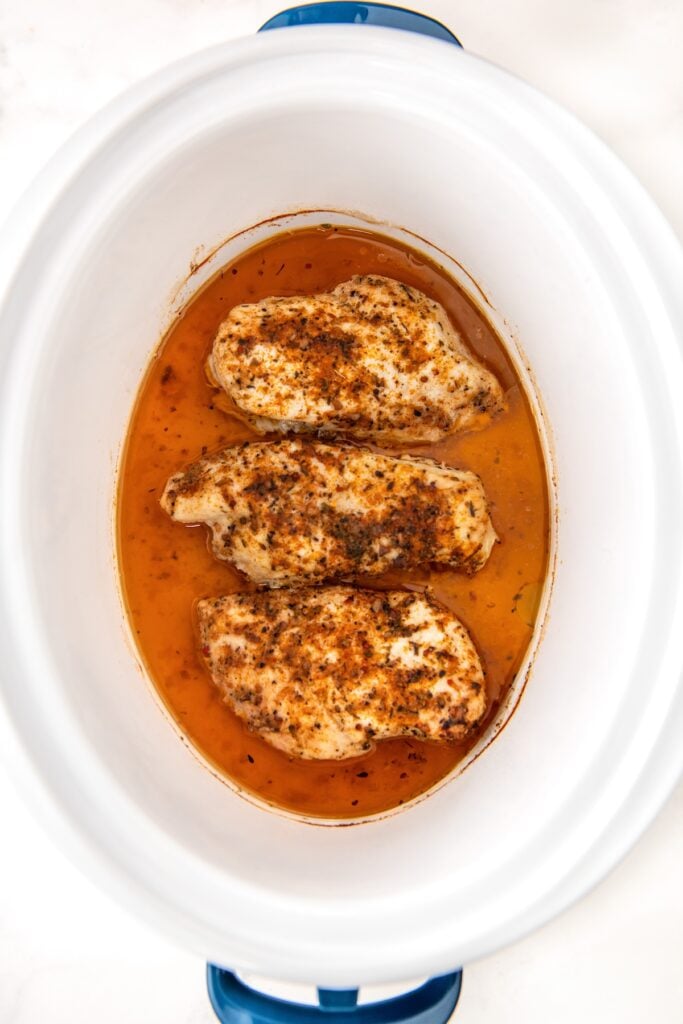 Three seasoned, cooked cajun chicken breasts sit in a white crockpot with orange-brown juices collected at the bottom.