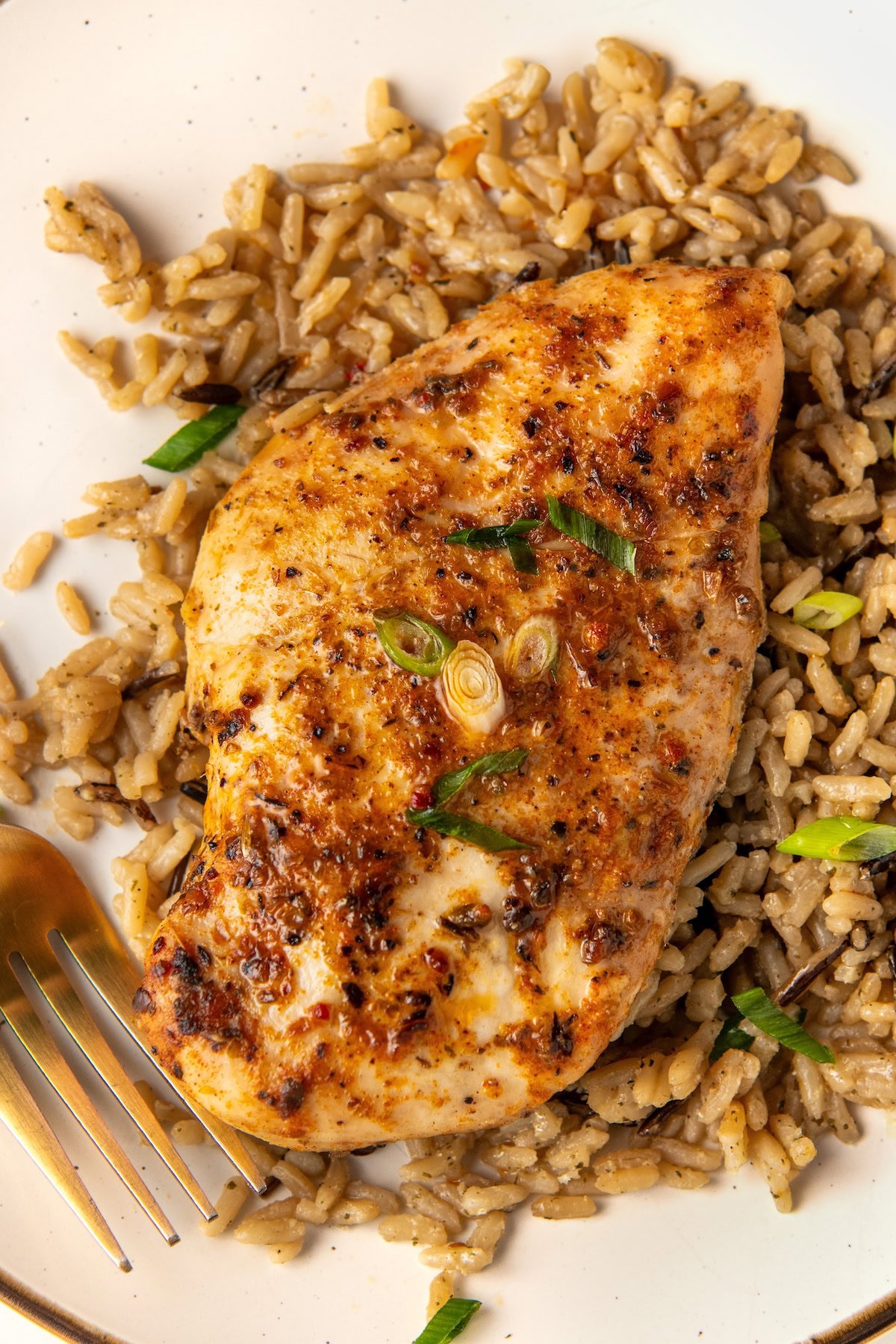 A seasoned, cooked crockpot cajun chicken breast sits atop a serving of dirty rice, garnished with chopped green onions, on a white plate with a fork.