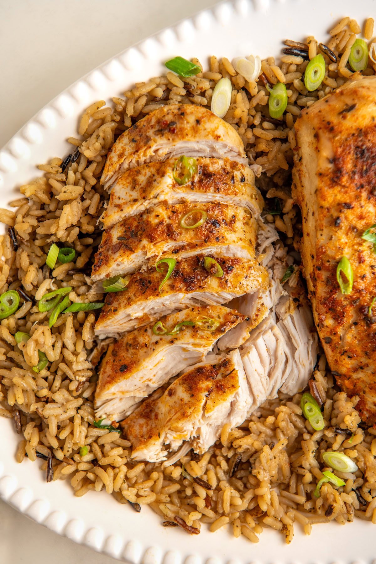 Sliced, seasoned crockpot cajun butter chicken is served on a bed of rice, garnished with chopped green onions on a white plate.
