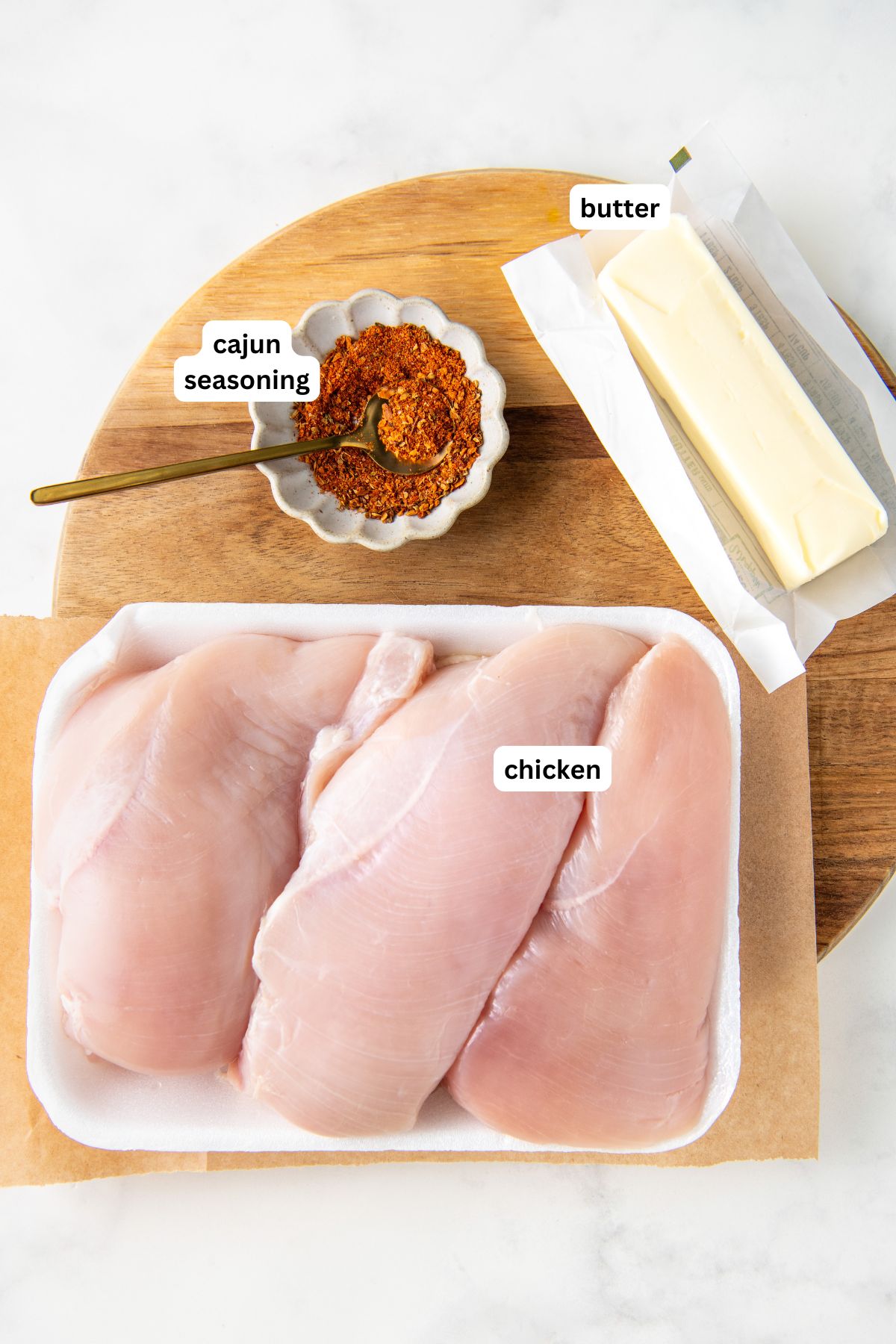 Ingredients for crockpot cajun butter chicken: raw chicken breasts on a tray, a bowl of Cajun seasoning with a spoon, and a stick of butter on a wooden board.