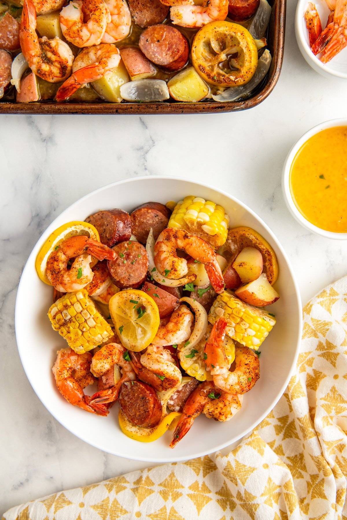 A white bowl filled with shrimp, sausage slices, corn on the cob pieces, potatoes, and lemon slices, with a sheet pan filled with a shrimp boil and a small bowl of butter sauce nearby on a marble surface.