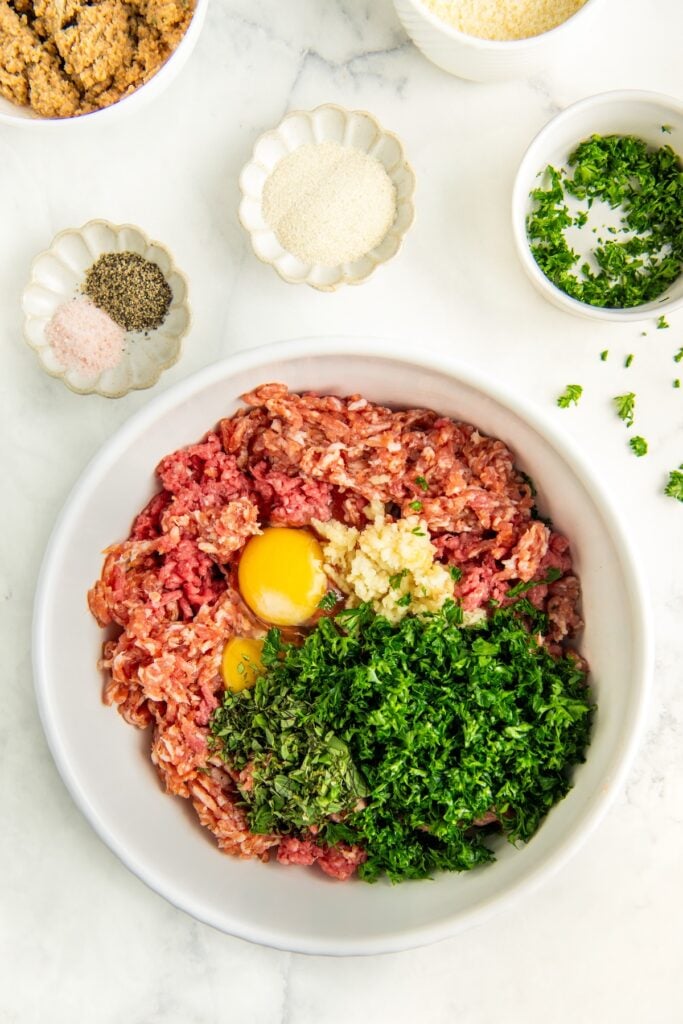 A white bowl contains ground meat, two eggs, chopped parsley, and minced garlic. Surrounding the bowl are small dishes with milk-soaked breadcrumbs, grated cheese, salt, pepper, and more chopped parsley on a white marble surface.