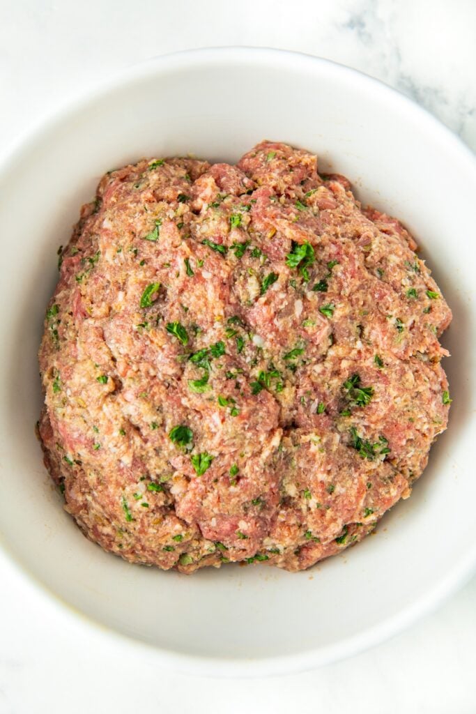 A white bowl filled with a mixture of raw meatball mixture blended together with herbs and seasonings, sitting on a white marble surface.