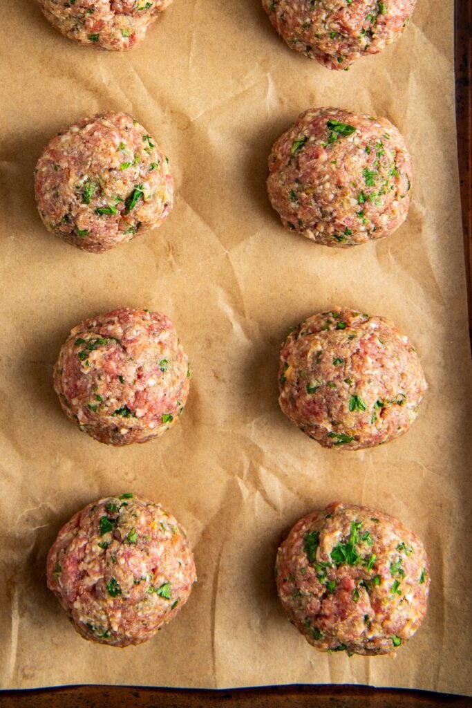 Eight raw homemade meatballs mixed with herbs are arranged on a baking sheet lined with brown parchment paper, ready for baking.