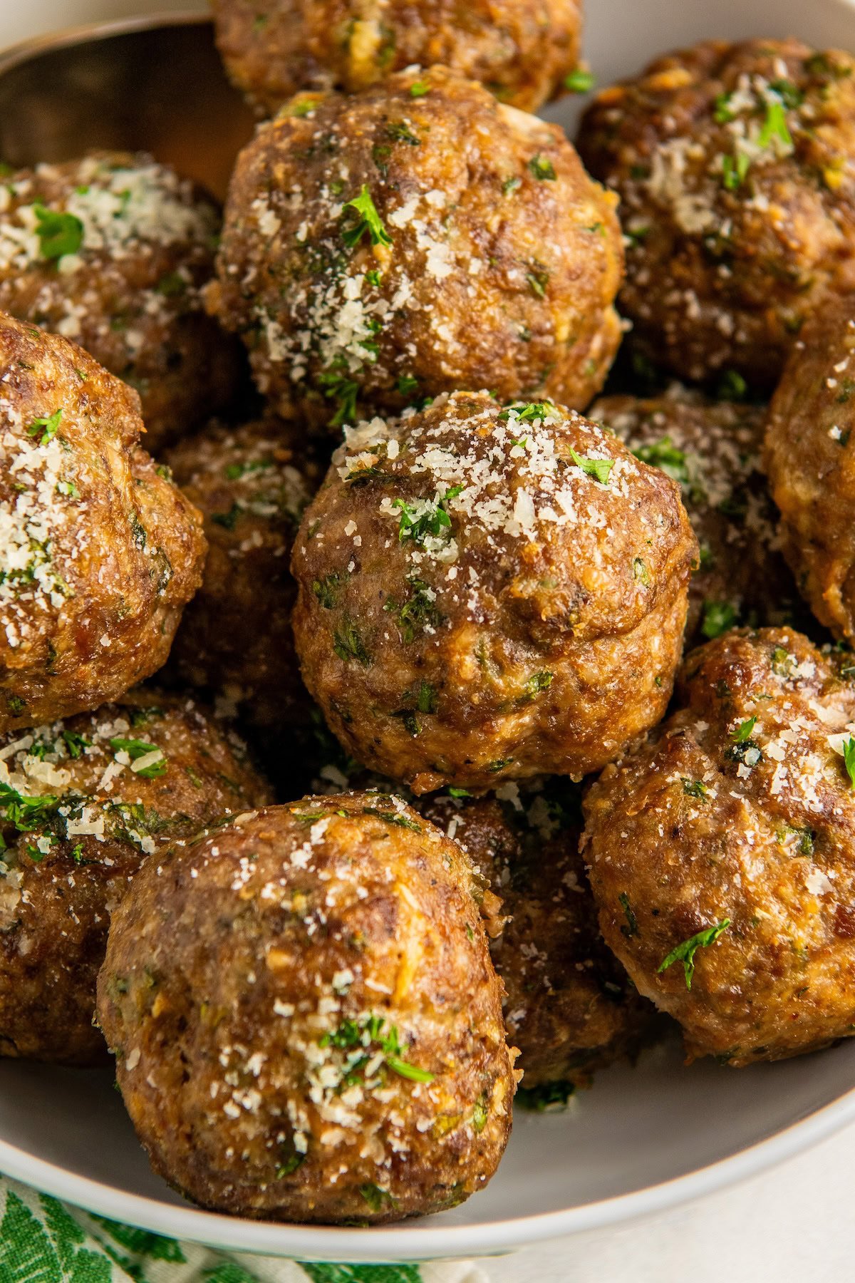 A close-up of several golden-brown baked Italian meatballs garnished with chopped parsley and grated Parmesan cheese, arranged in a white bowl.