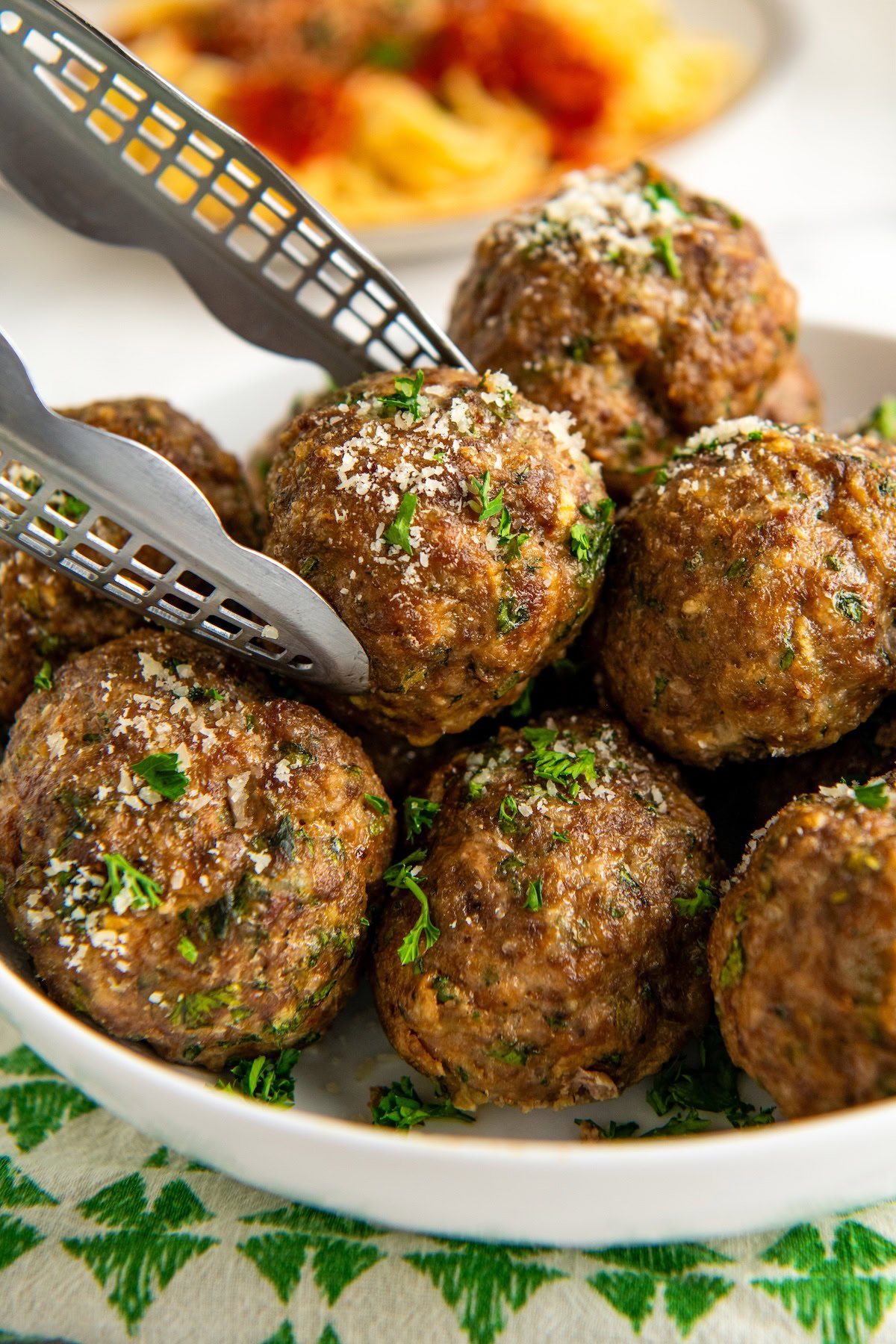 A close-up of metal tongs holding a large, seasoned homemade meatball topped with grated cheese and chopped parsley, with more meatballs arranged in a white bowl. A blurred plate of spaghetti pasta is visible in the background.