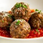 Four baked Italian meatballs garnished with chopped parsley and grated cheese sit on a bed of tomato sauce in a white bowl.