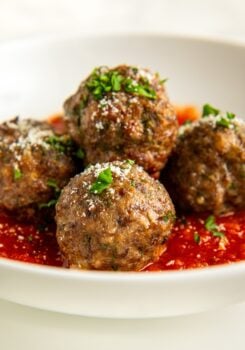 Four baked Italian meatballs garnished with chopped parsley and grated cheese sit on a bed of tomato sauce in a white bowl.