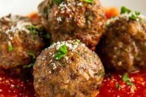 Four baked Italian meatballs garnished with chopped parsley and grated cheese sit on a bed of tomato sauce in a white bowl.
