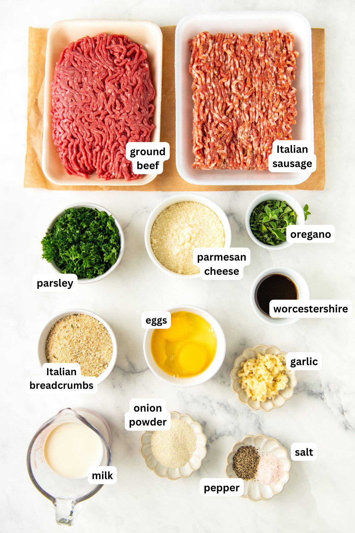 Top-down view of ingredients for this Italian meatball recipe arranged on a white surface, including ground beef, Italian ground sausage, parsley, oregano, parmesan cheese, Worcestershire sauce, eggs, breadcrumbs, garlic, milk, onion powder, salt, and pepper.