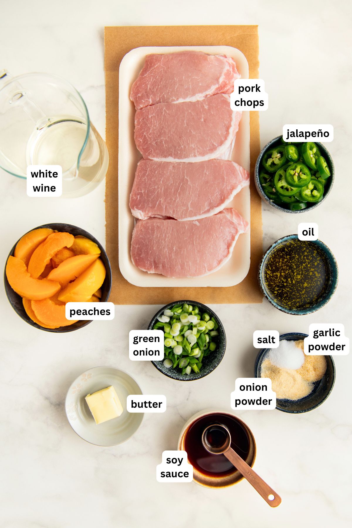 Recipe ingredients arranged in bowls for jalapeño peach pork chop recipe showing raw pork chops, sliced jalapeño, green onion, butter, white wine, peaches, soy sauce, oil, salt, garlic powder, and onion powder, each ingredient labeled on a white background.