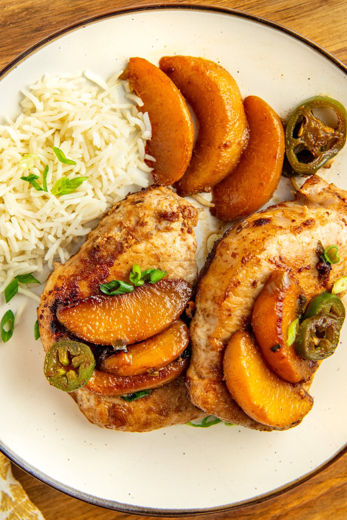 A plate with two seared pork chops topped with cooked peach slices and jalapeño slices, served with white rice and garnished with chopped green onions.