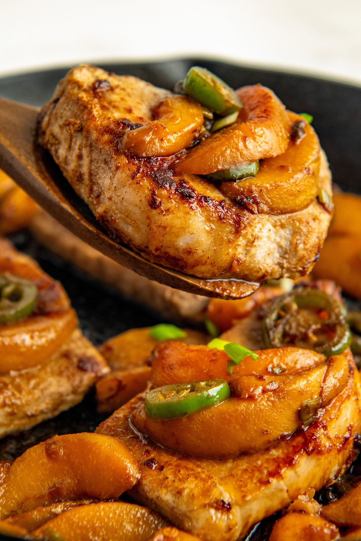 A close-up of a seared pork chop topped with sliced peaches and jalapeños, lifted by a wooden spatula. More pork chops with the same toppings are visible in the skillet below.