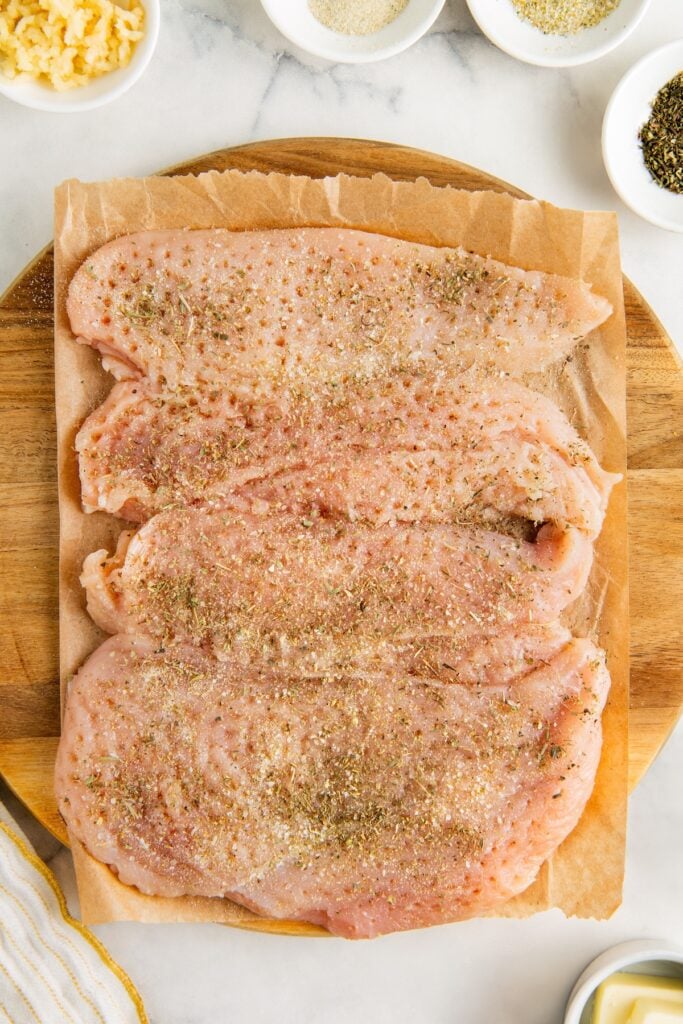 Three raw, tenderized and seasoned chicken breasts are placed on parchment paper atop a wooden board, with small bowls of minced garlic, spices, and herbs around them on a white surface.