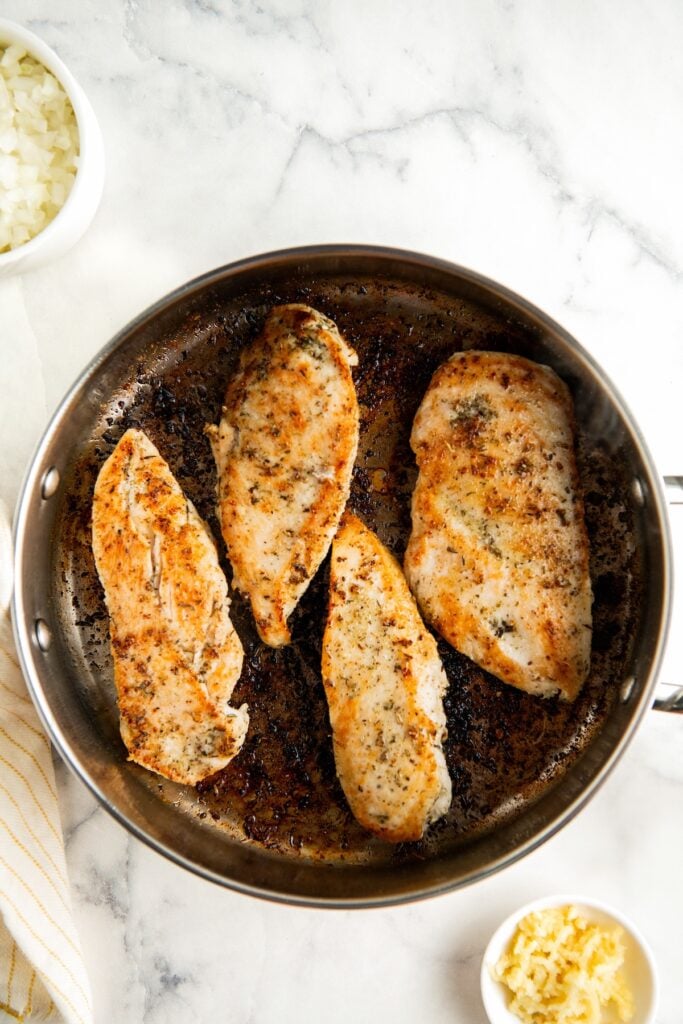 Four seasoned chicken breasts are cooking in a stainless steel skillet. The chicken is browned and seared. Small bowls of chopped onions and minced garlic are nearby on a white marble surface.