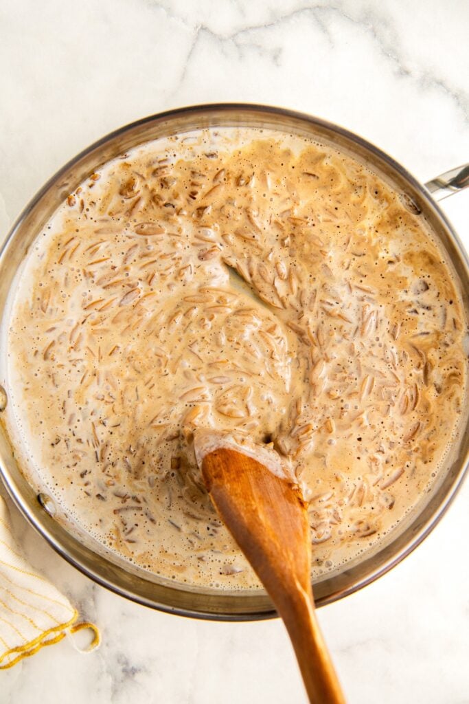 A saucepan filled with orzo pasta cooking in a creamy lemon sauce, being stirred with a wooden spoon on a marble countertop.
