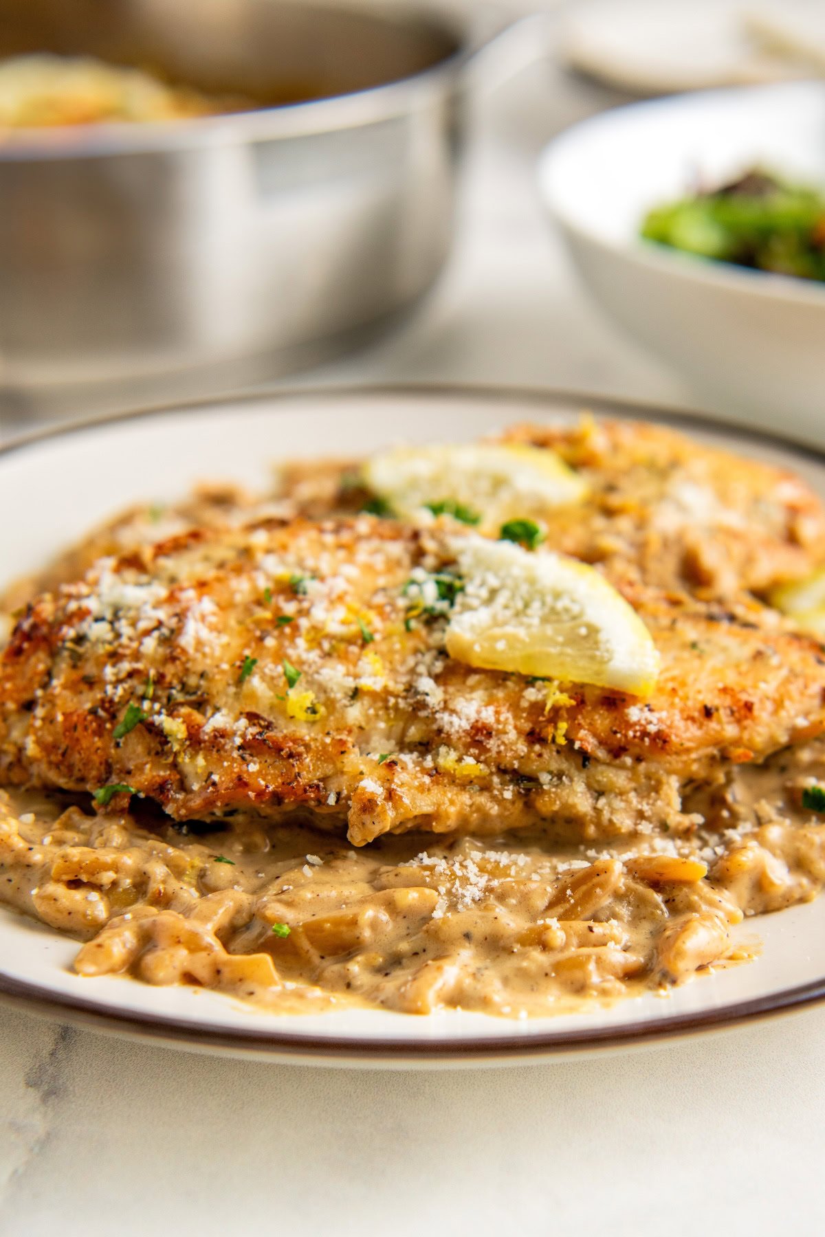 A plate of creamy lemon chicken orzo garnished with lemon slices, parsley, and grated cheese. A pot and a bowl of salad are blurred in the background.