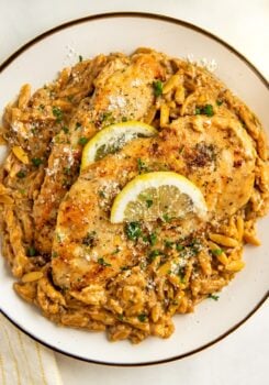 A plate of creamy lemon orzo pasta topped with two golden-brown crispy chicken breasts, garnished with lemon slices, fresh herbs, and grated cheese.