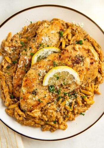 A plate of creamy lemon orzo pasta topped with two golden-brown crispy chicken breasts, garnished with lemon slices, fresh herbs, and grated cheese.