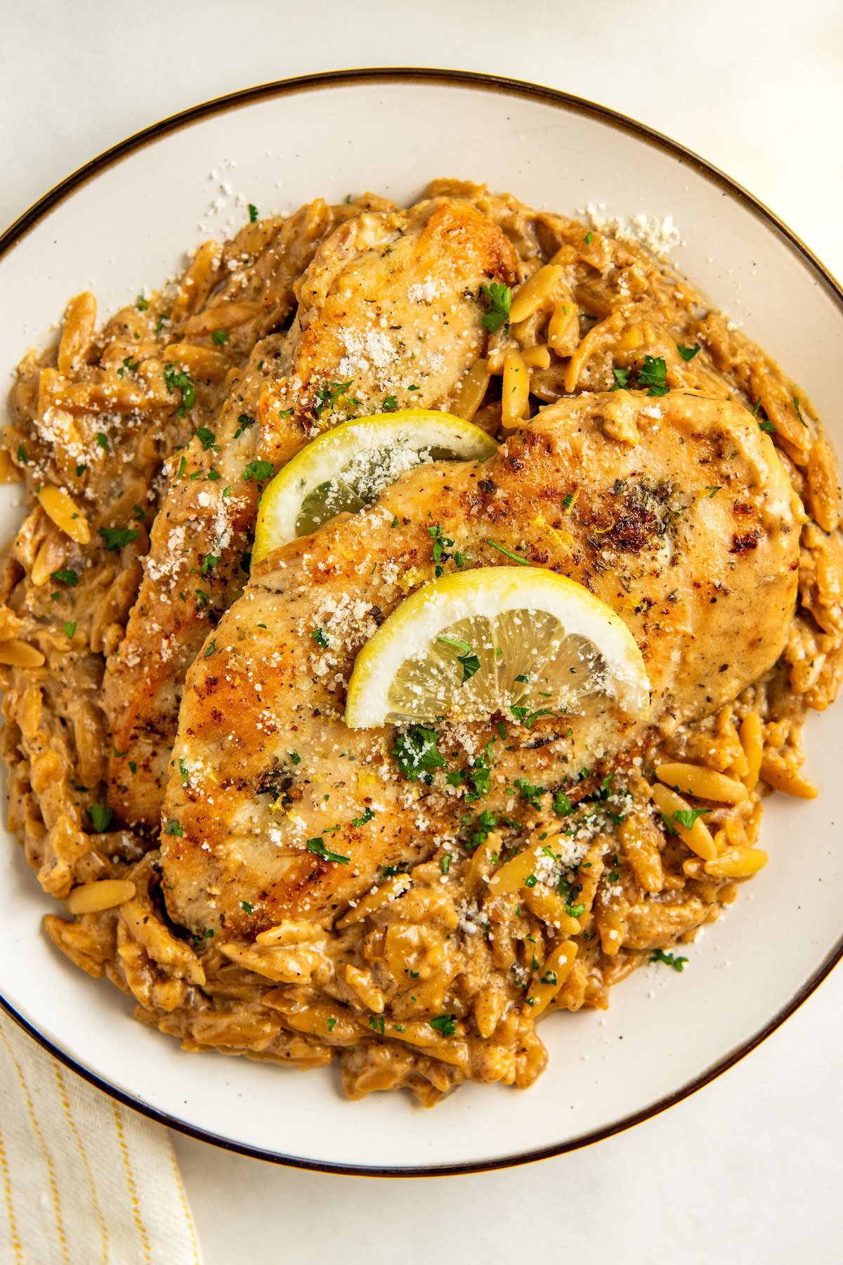 Ready in 30 minutes, this easy Lemon Orzo Chicken recipe features juicy chicken and tender orzo coated in a rich and creamy lemon sauce. A plate of creamy lemon orzo pasta topped with two golden-brown crispy chicken breasts, garnished with lemon slices, fresh herbs, and grated cheese.