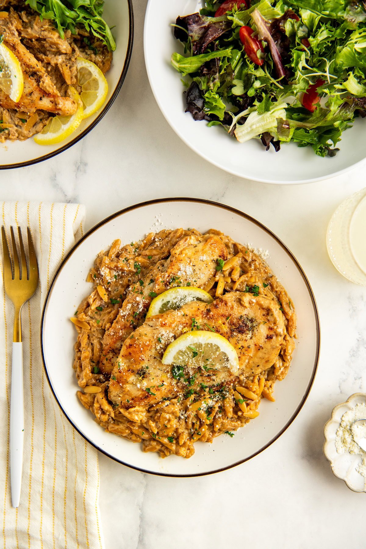 A plate of creamy lemon chicken orzo pasta topped with lemon slices and sprinkled with herbs and cheese, beside a green salad and a yellow-striped napkin with a gold fork.