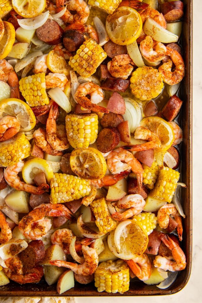 A sheet pan filled with cooked shrimp, sliced sausage, corn on the cob, potato chunks, onions, and lemon slices, all seasoned and roasted.