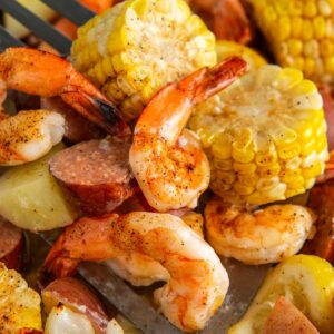 A close-up of a sheet pan shrimp boil with a spatula scooping up seasoned shrimp, sliced sausage, corn on the cob, and chunks of potato.