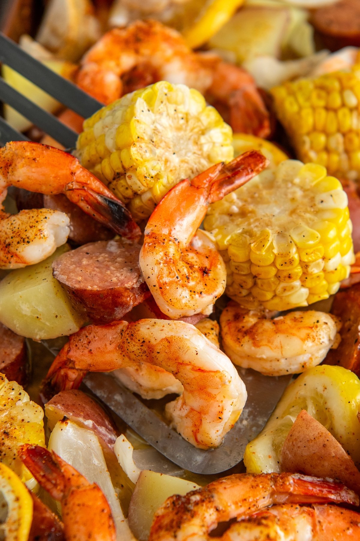 A close-up of a sheet pan shrimp boil with a spatula scooping up seasoned shrimp, sliced sausage, corn on the cob, and chunks of potato.