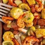 A sheet pan shrimp boil filled with seasoned baked shrimp, corn on the cob slices, red potatoes, sausage, lemon wedges, and chopped herbs, with a spatula lifting a serving.